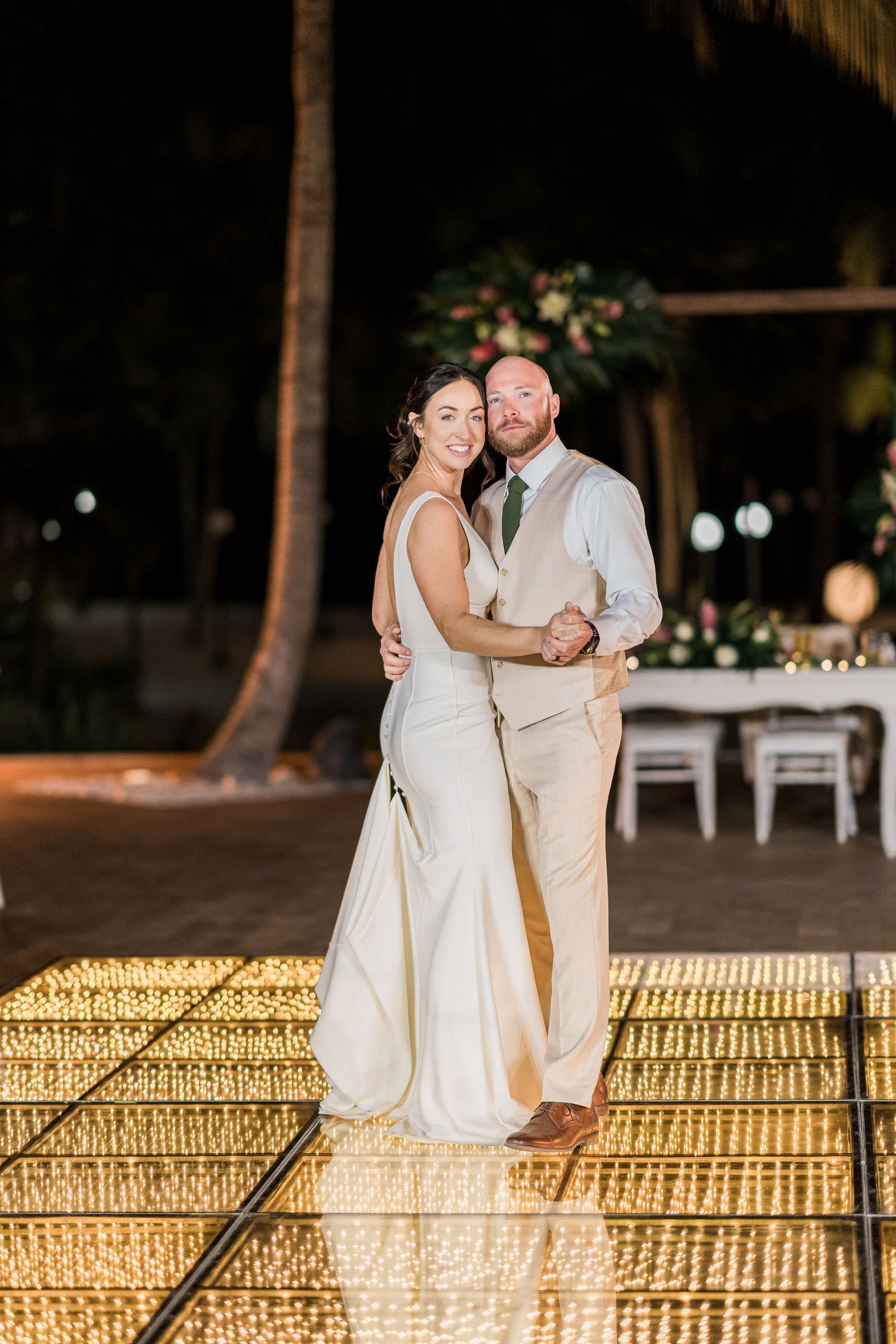 Bride and groom share first dance on glowing LED dance floor at outdoor tropical wedding reception at night.