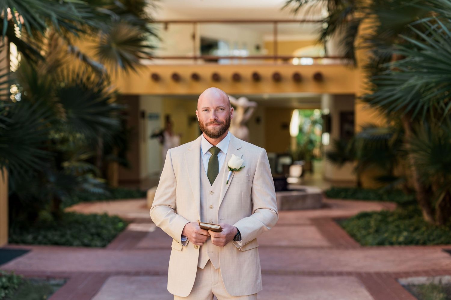 Bald groom in beige suit with white boutonniere and green tie poses in tropical courtyard.