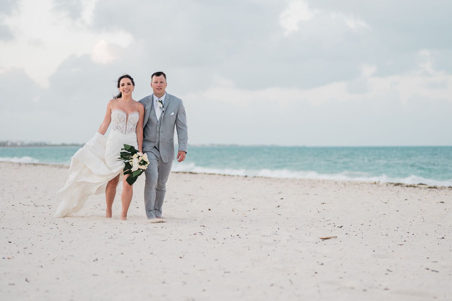 Bride and groom walking hand in hand on a white sand beach with turquoise ocean waves in the background.