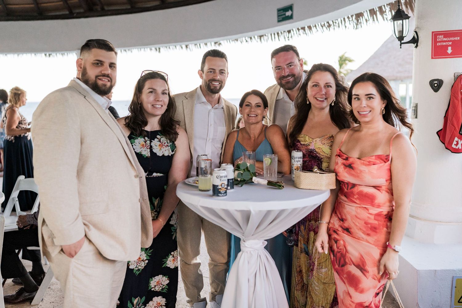 Group of seven friends posing together at an outdoor wedding reception venue with tropical decor.