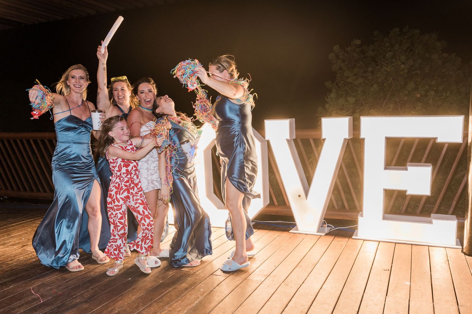 Group of women in elegant gowns posing joyfully beside illuminated LOVE letters on a wooden deck at a wedding reception.