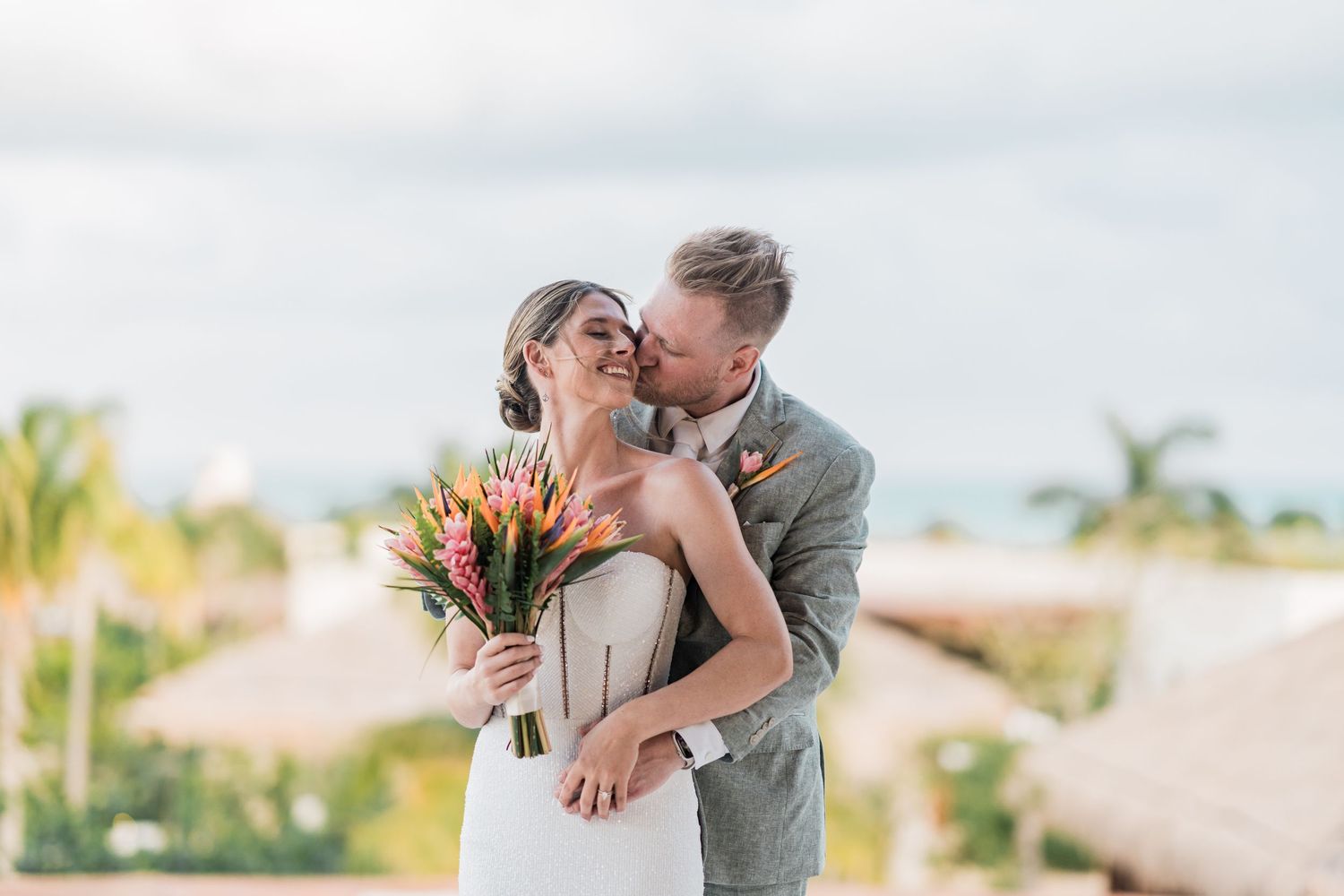 Bride and groom share a romantic kiss outdoors, bride holding vibrant tropical bouquet at a destination wedding.