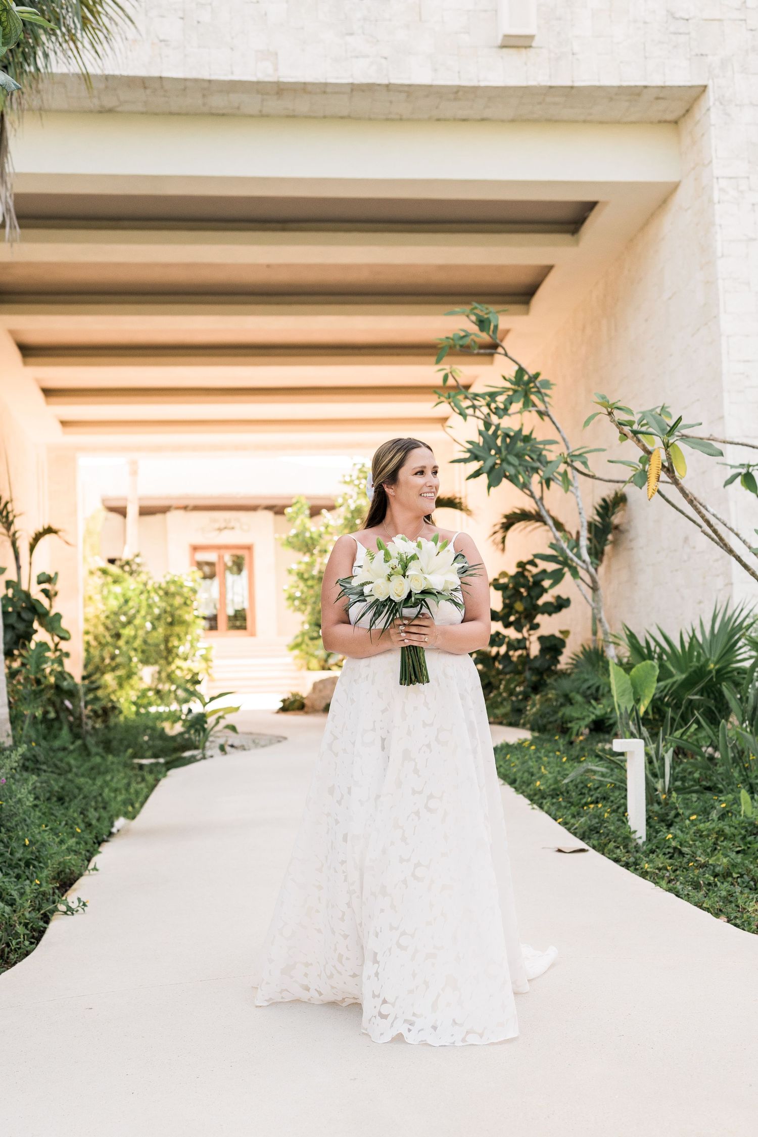 Bride in lace wedding gown holding white bouquet walks along tropical garden pathway at luxury resort venue.