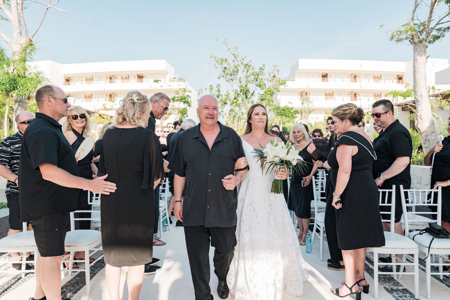Bride walks down outdoor wedding aisle with escort, guests in black attire, white chairs, tropical resort backdrop.