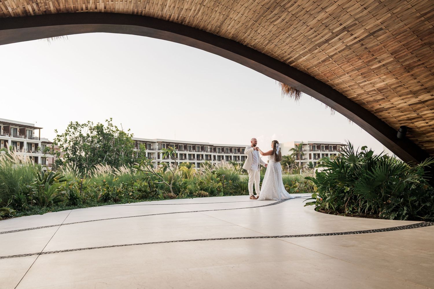 Couple in white attire holding hands at a luxury resort, framed by an elegant arch with lush greenery.