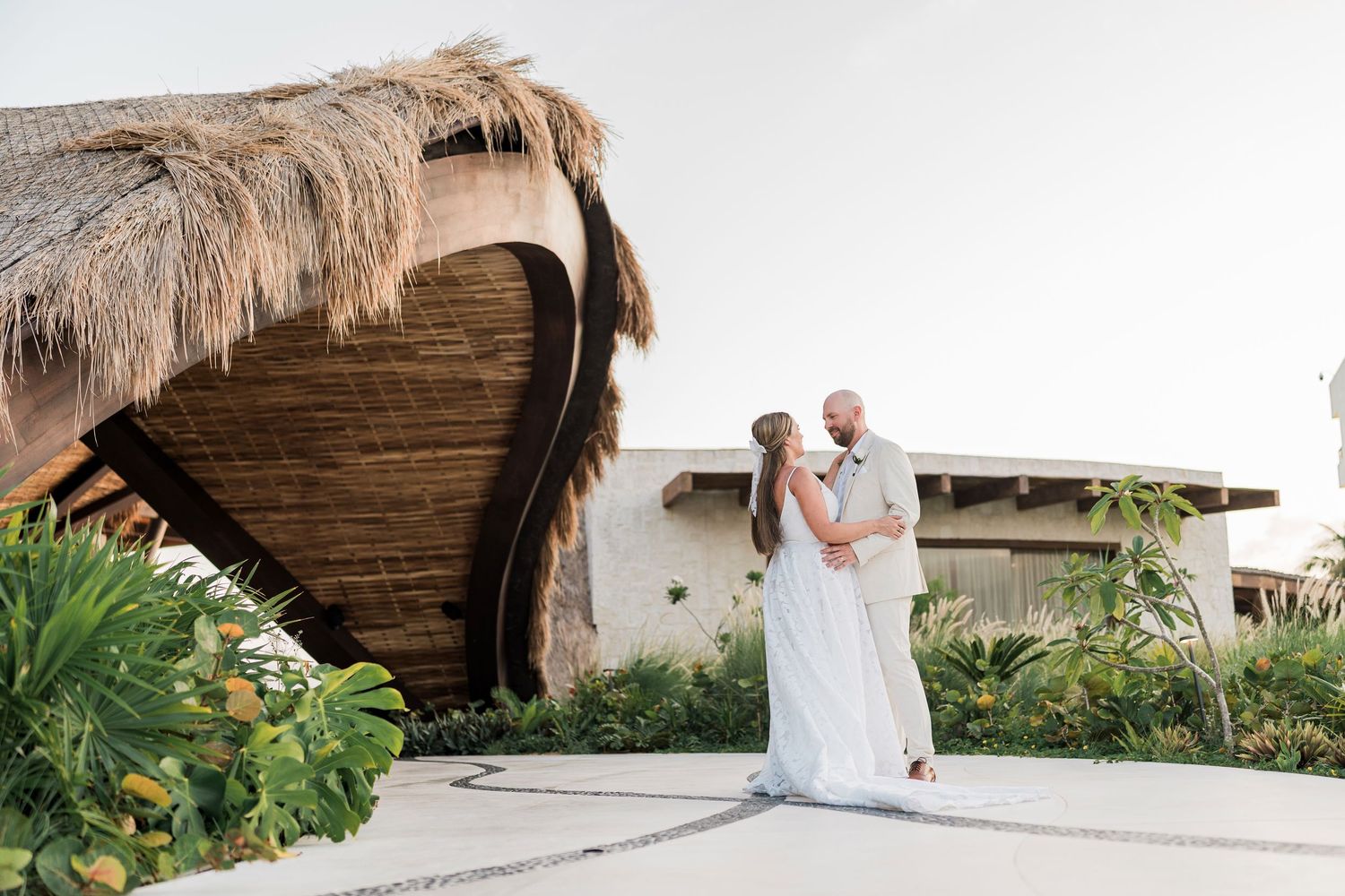 Couple shares romantic embrace in white wedding attire outside a tropical resort with thatched roof architecture.