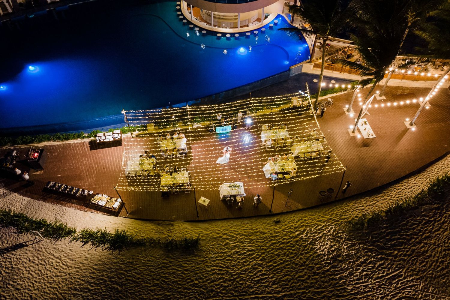 Aerial night view of an elegant beachside wedding reception with string lights and decorated tables near a pool. Puerto Vallarta wedding