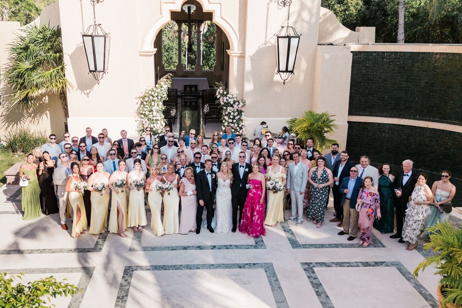 Large wedding group photo outside elegant white villa with bride in white, groom, bridesmaids in gold, and guests.