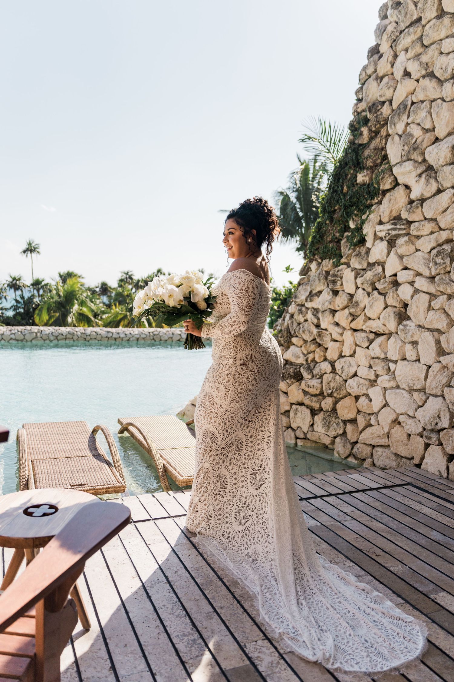 Xcaret Arte Wedding - Bride in lace long-sleeve wedding gown holding white bouquet on tropical resort deck by stone wall.