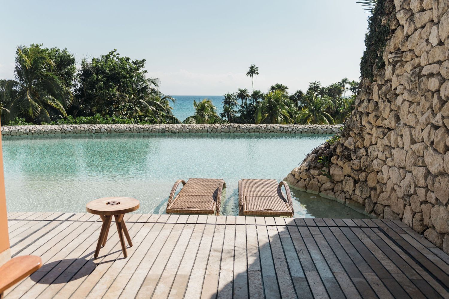Xcaret Arte Wedding - Luxury pool with wooden deck, lounge chairs, and ocean view surrounded by tropical palms and stone wall.