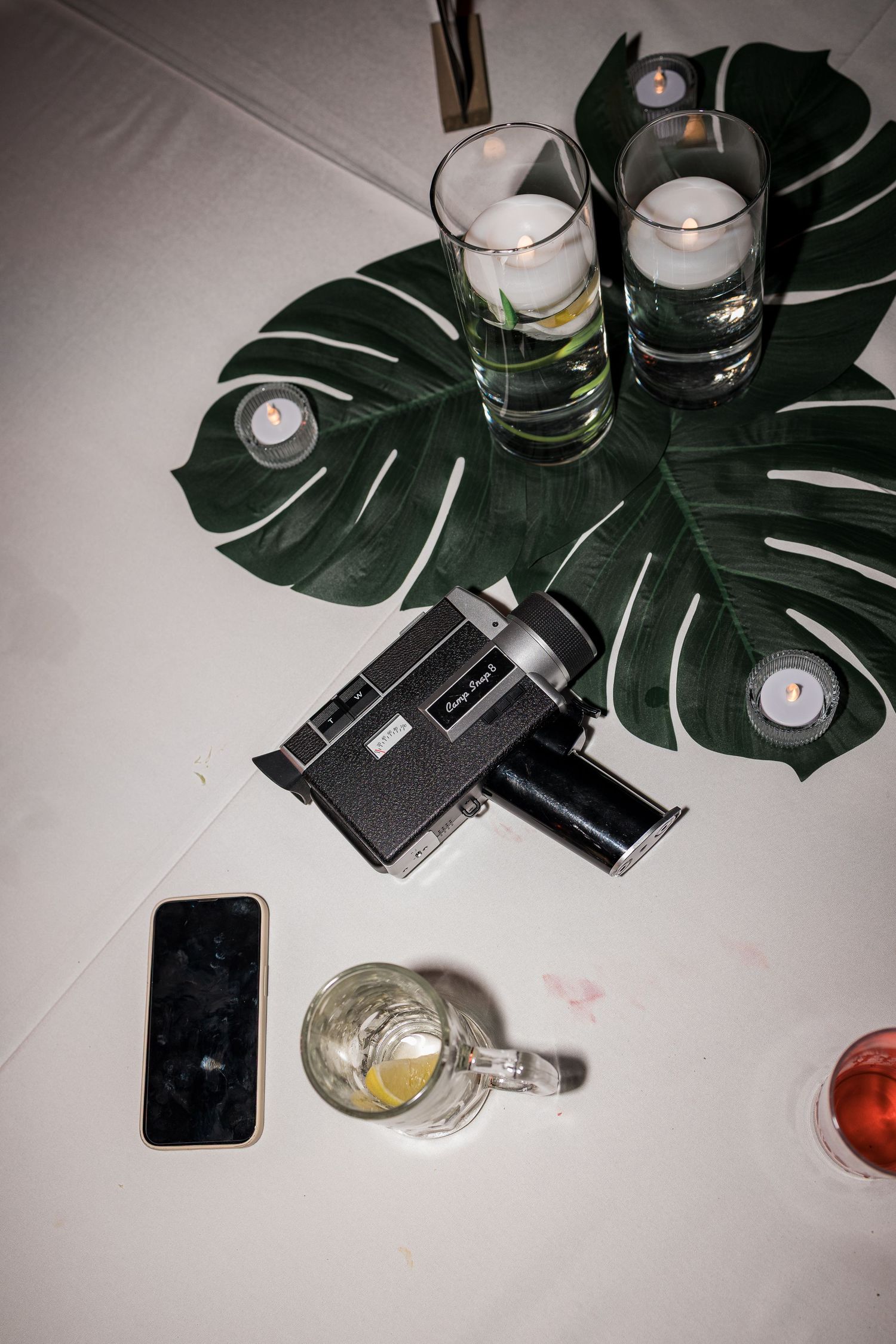 Vintage camera, smartphone, candles, and monstera leaf centerpiece on white table at elegant event.