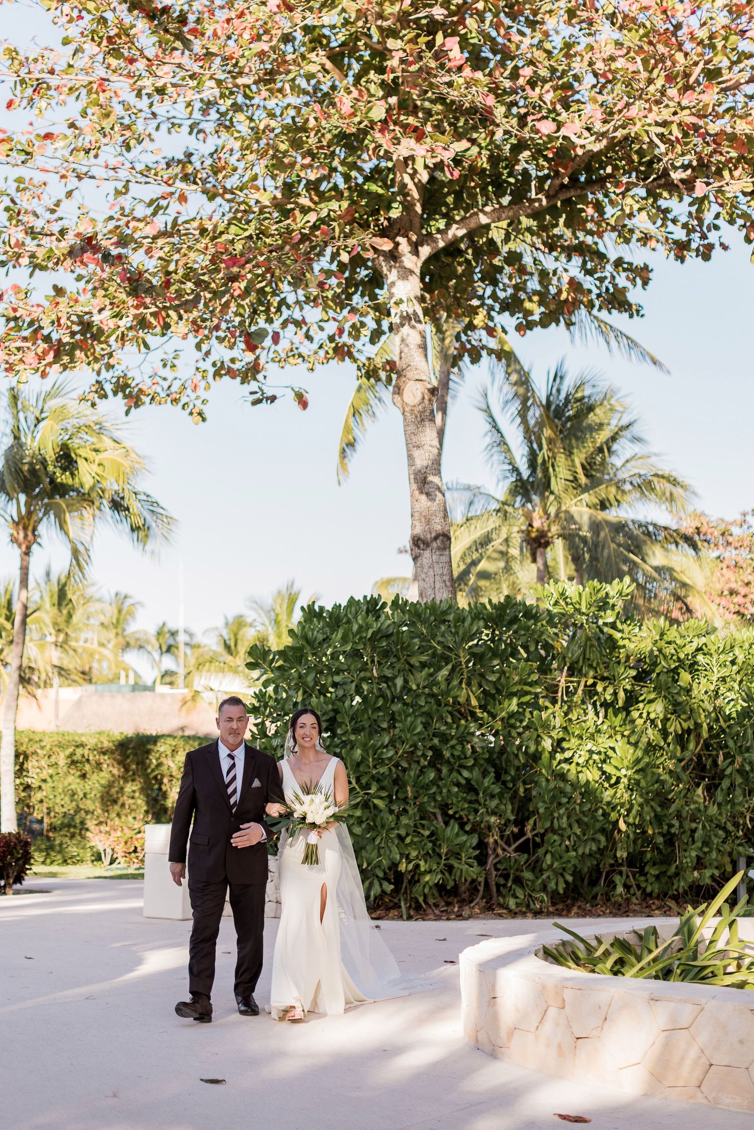 Bride and groom walk together outdoors under a colorful tree with palm trees and lush greenery at a tropical wedding venue.