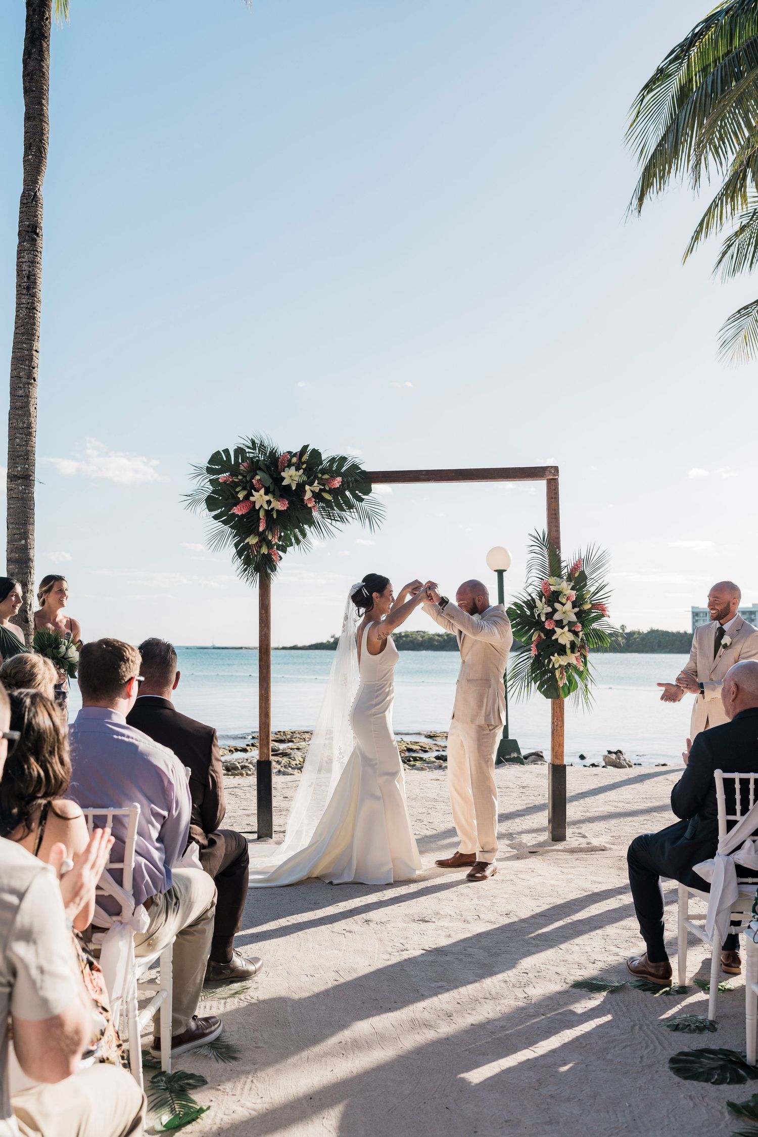 Bride and groom hold hands under floral arch during beachside wedding ceremony with guests seated on white chairs.