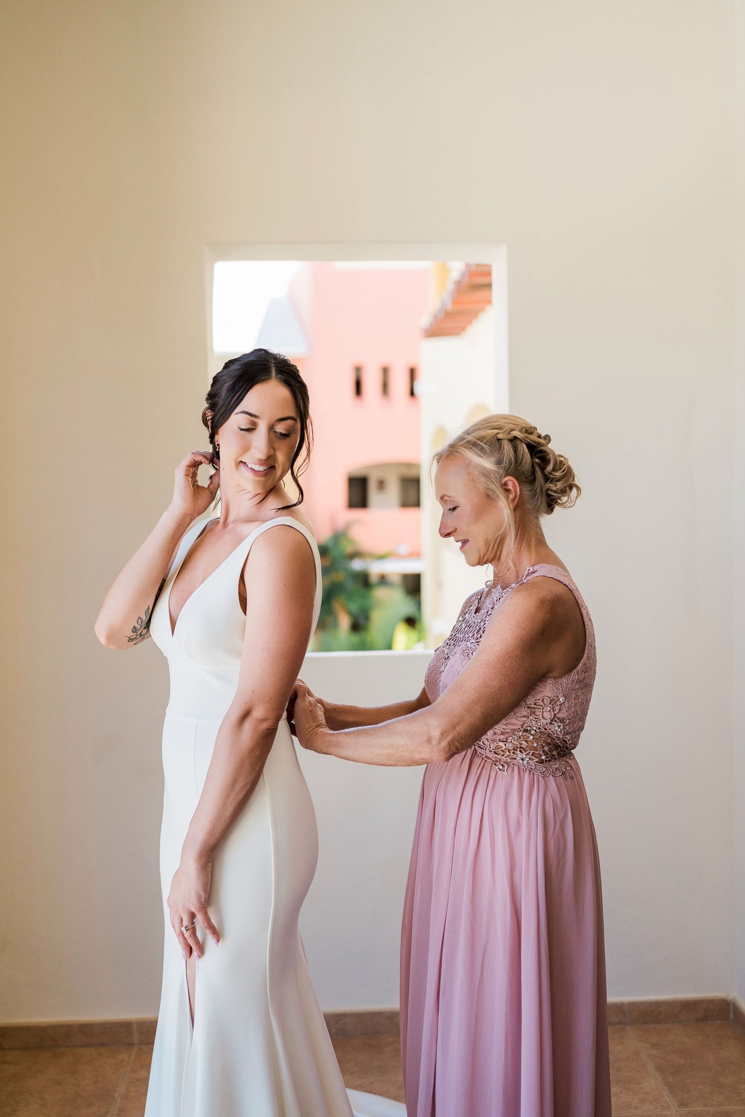 Bride in white gown smiles as woman in pink dress helps fasten her wedding dress near a sunlit window.