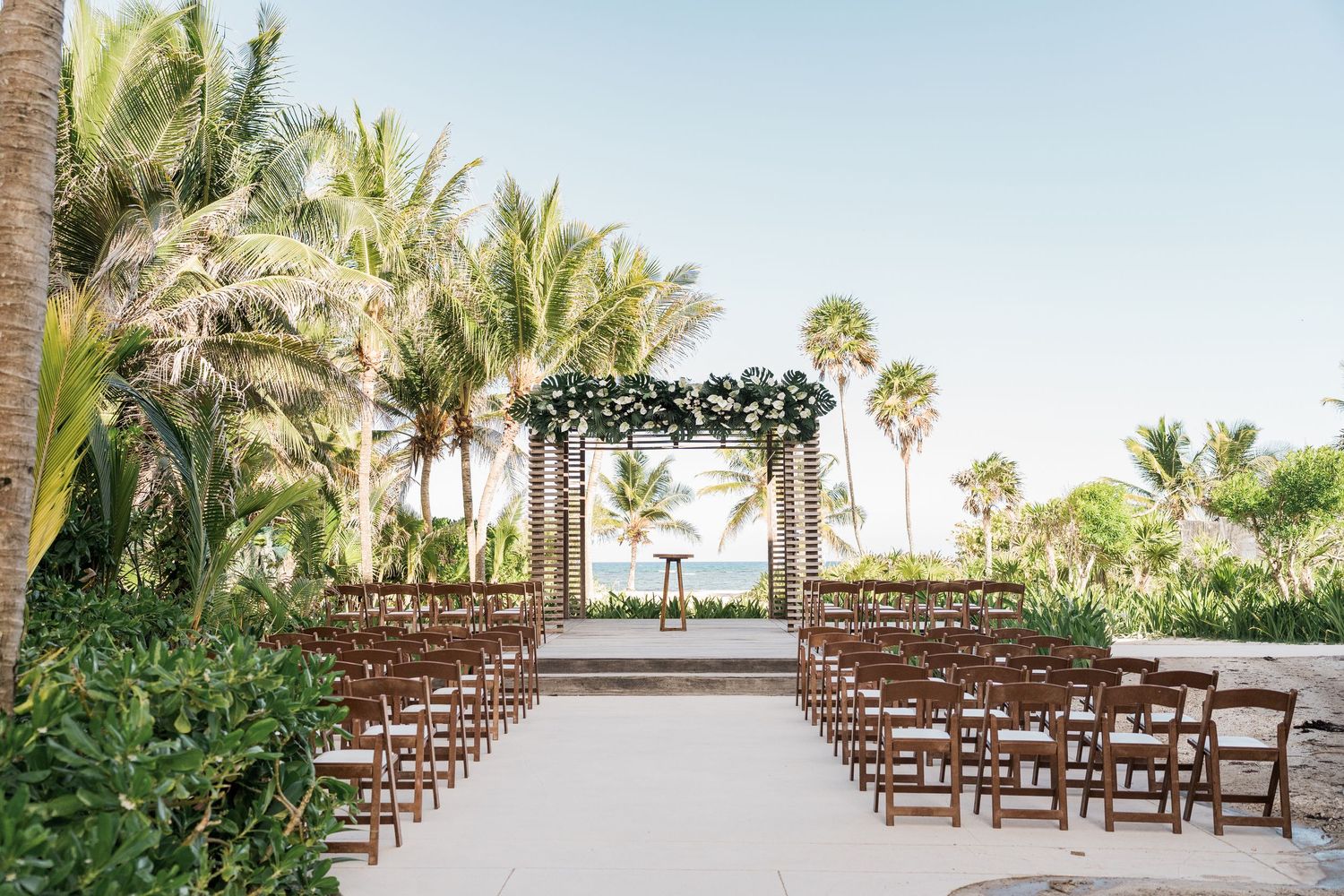 Elegant outdoor beach wedding ceremony setup with floral arch and palm trees.