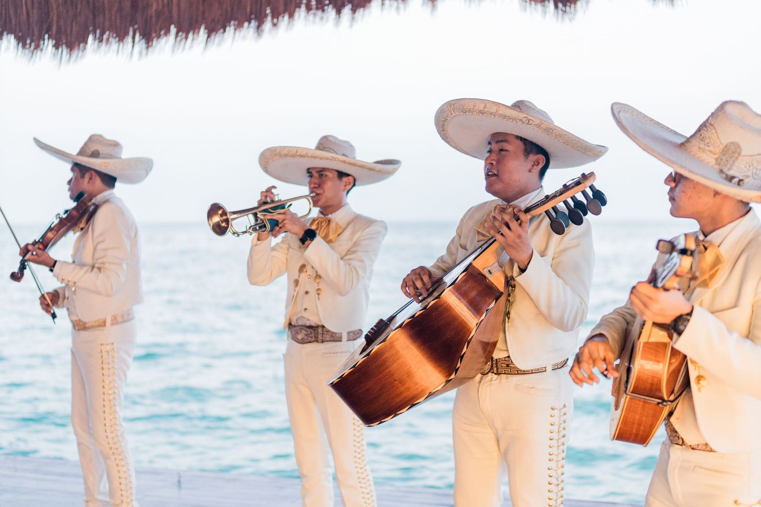 Mariachi band in white charro suits playing instruments on a beach at a destination wedding celebration.