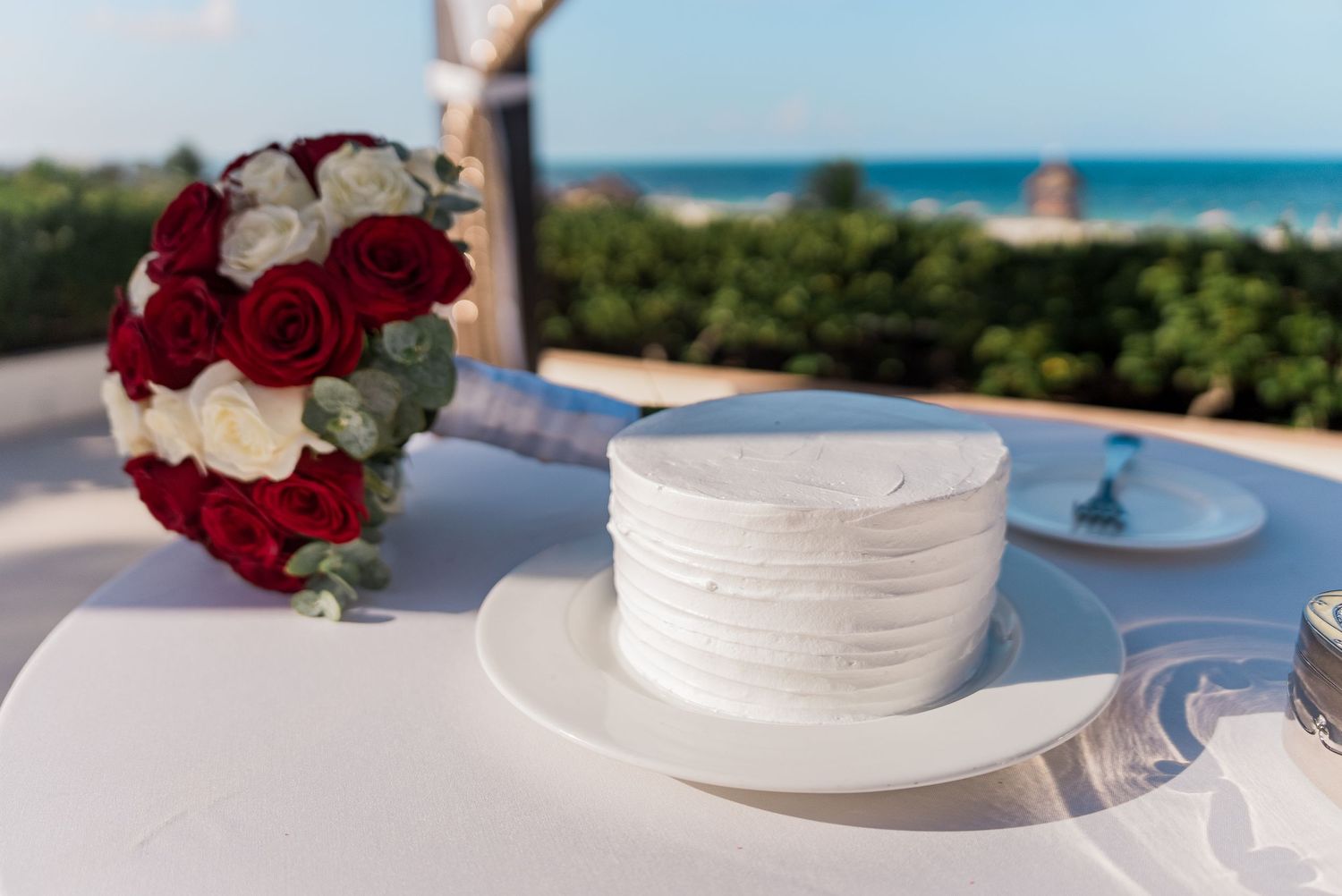 White wedding cake beside red and white rose bridal bouquet with ocean view.