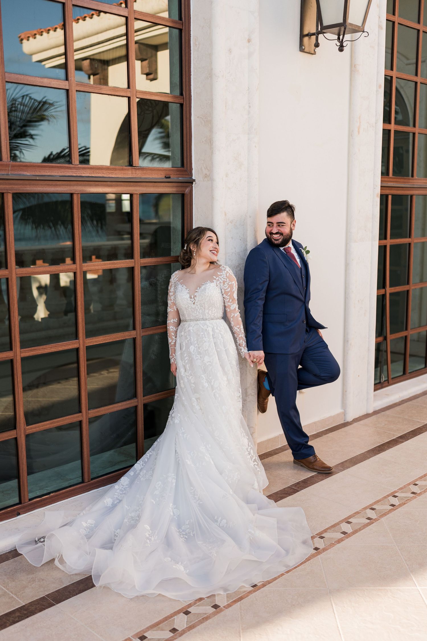 Bride in lace gown and groom in navy suit hold hands smiling outside elegant venue.