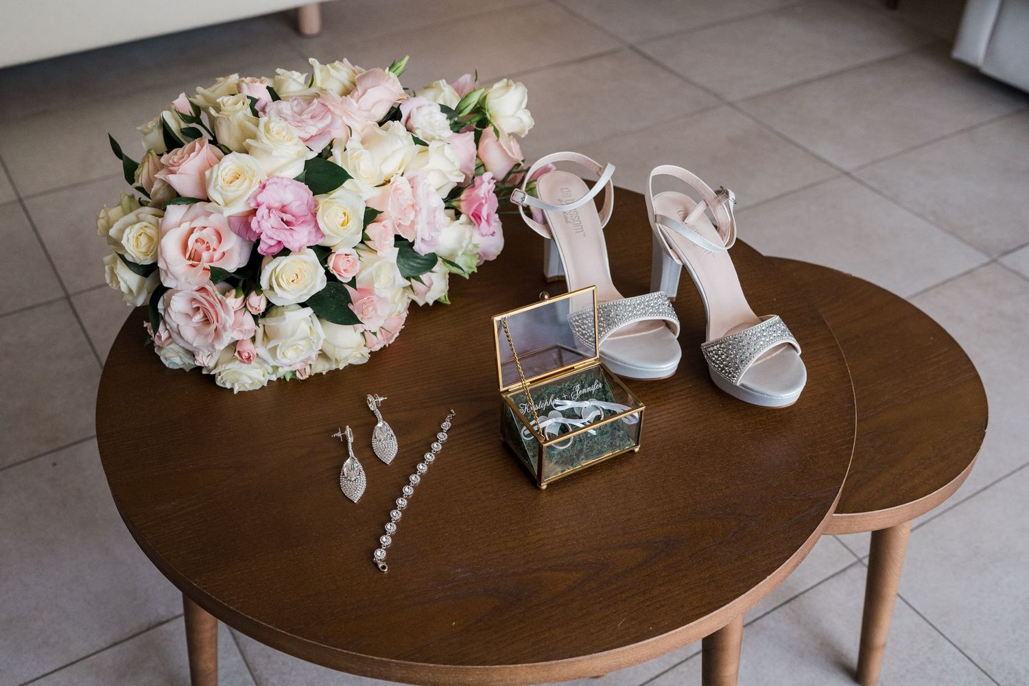 Bridal accessories including pink rose bouquet, heels, earrings, and jewelry box on table.