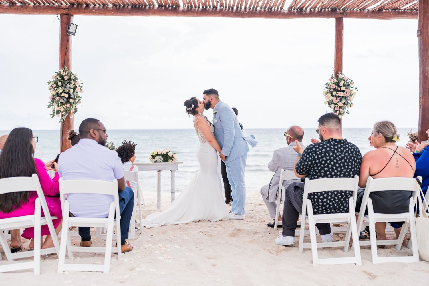 Newlyweds share first kiss at beach wedding ceremony while guests watch from white chairs on sand.