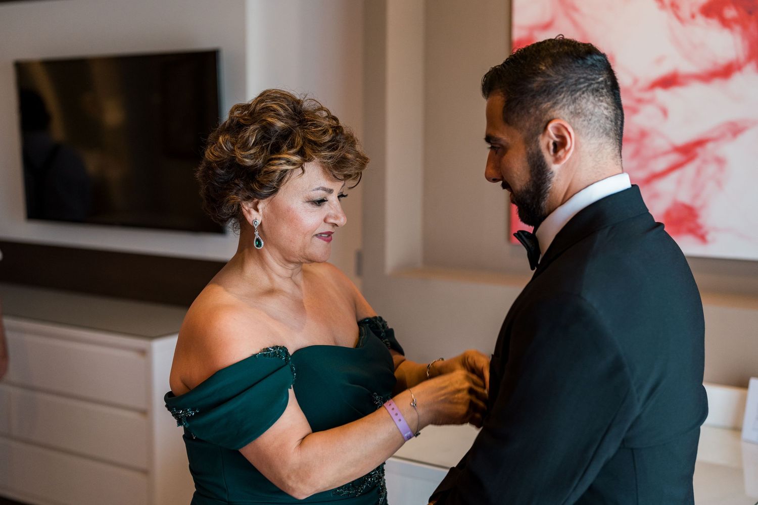Mother pins boutonniere on groom wearing black tuxedo before wedding ceremony.