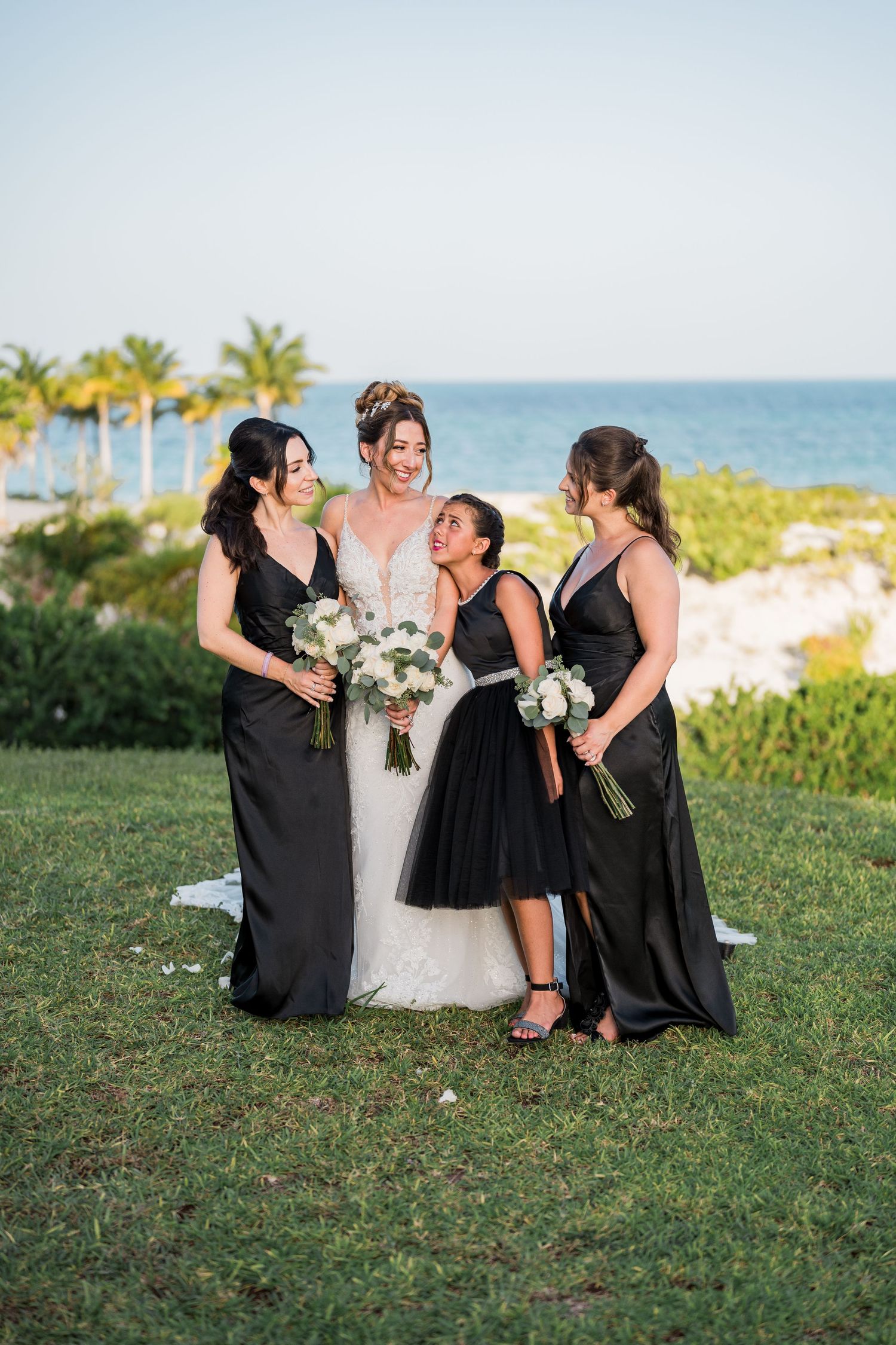 Bride in lace gown stands with three bridesmaids in black dresses holding white bouquets by the sea.