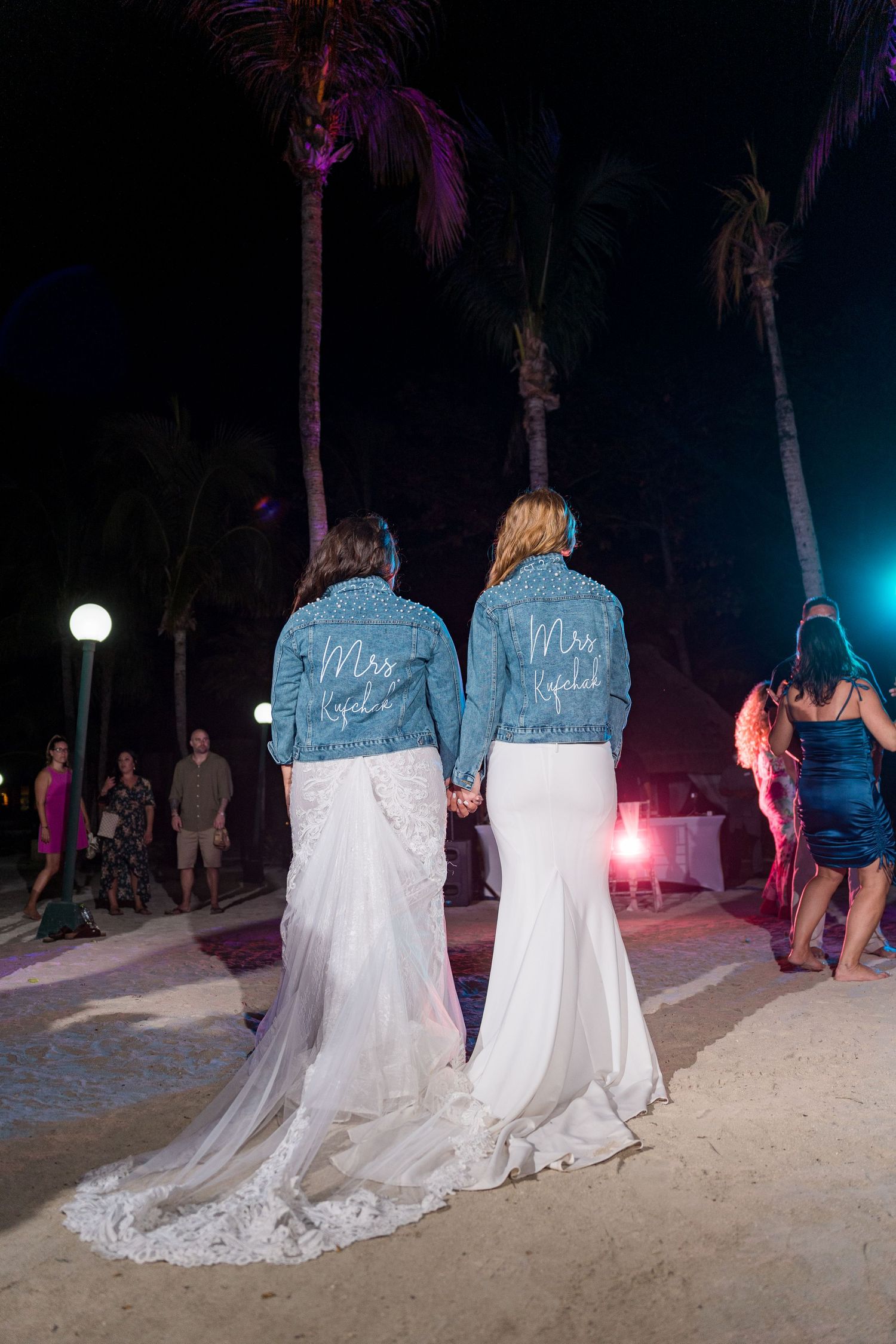 Two brides in white gowns and matching denim jackets holding hands on beach.
