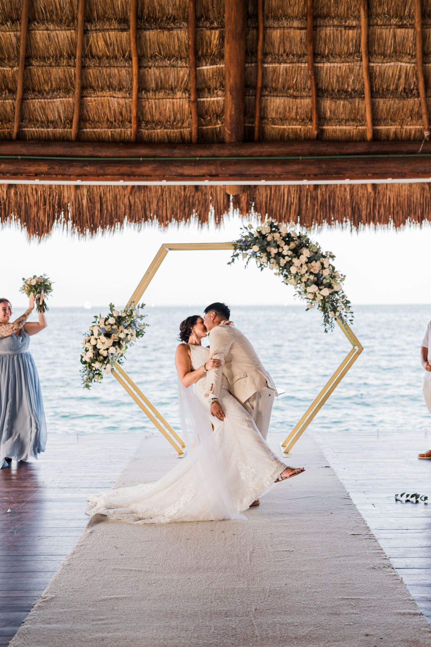 Newlyweds share first kiss under gold hexagonal arch at beachside wedding ceremony.