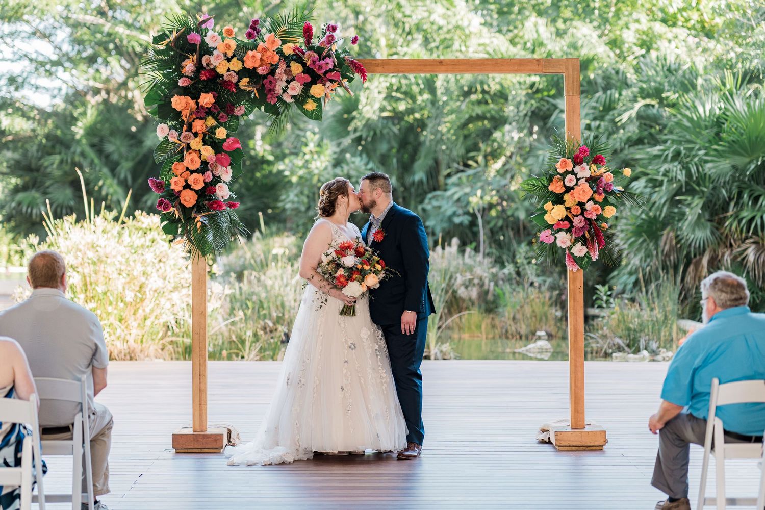 Secrets Akumal Wedding - Bride and groom share first kiss under a wooden arch adorned with colorful floral arrangements
