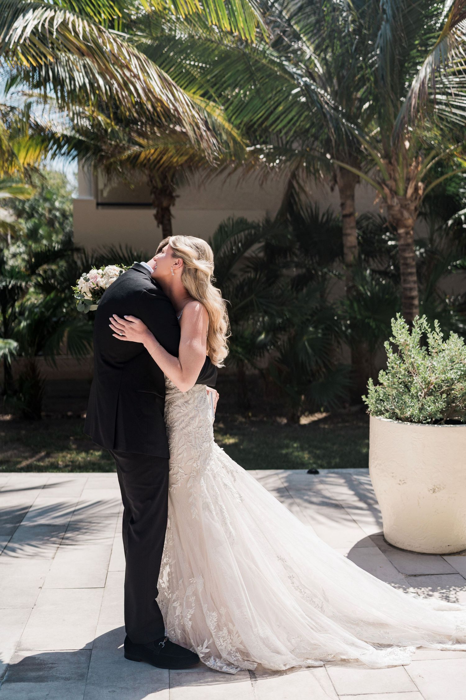 Bride and groom share a romantic embrace outdoors, surrounded by lush palm trees, as she holds a white bouquet in her lace gown.