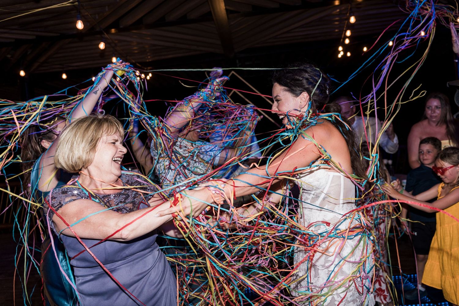 Bride and older woman dancing joyfully covered in colorful streamers at a wedding reception celebration.