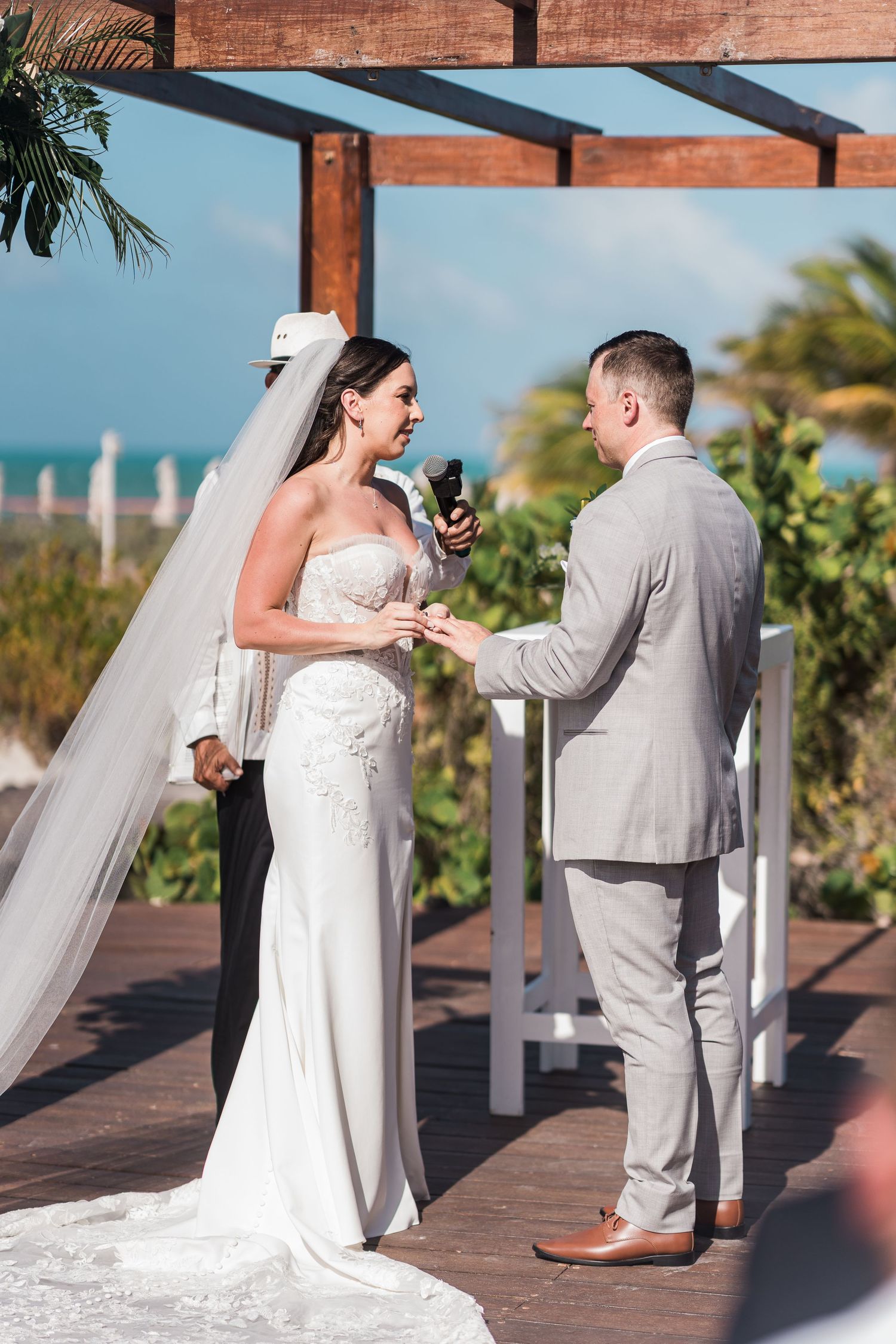 Bride and groom exchange rings during outdoor beach wedding ceremony under wooden pergola with ocean view.