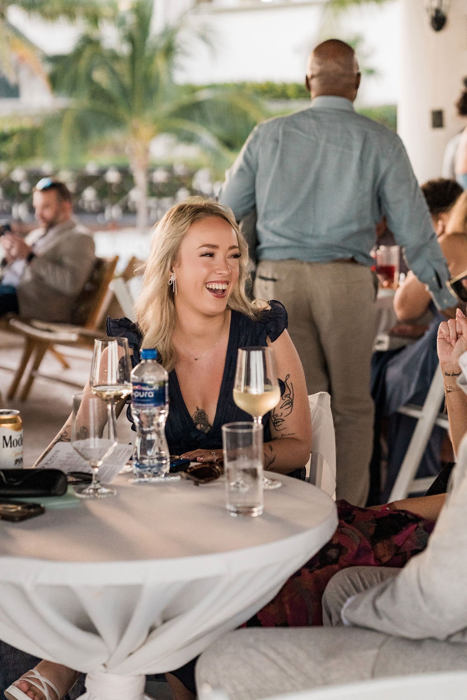 Laughing blonde woman with tattoos enjoying white wine at an outdoor restaurant table in a tropical setting.