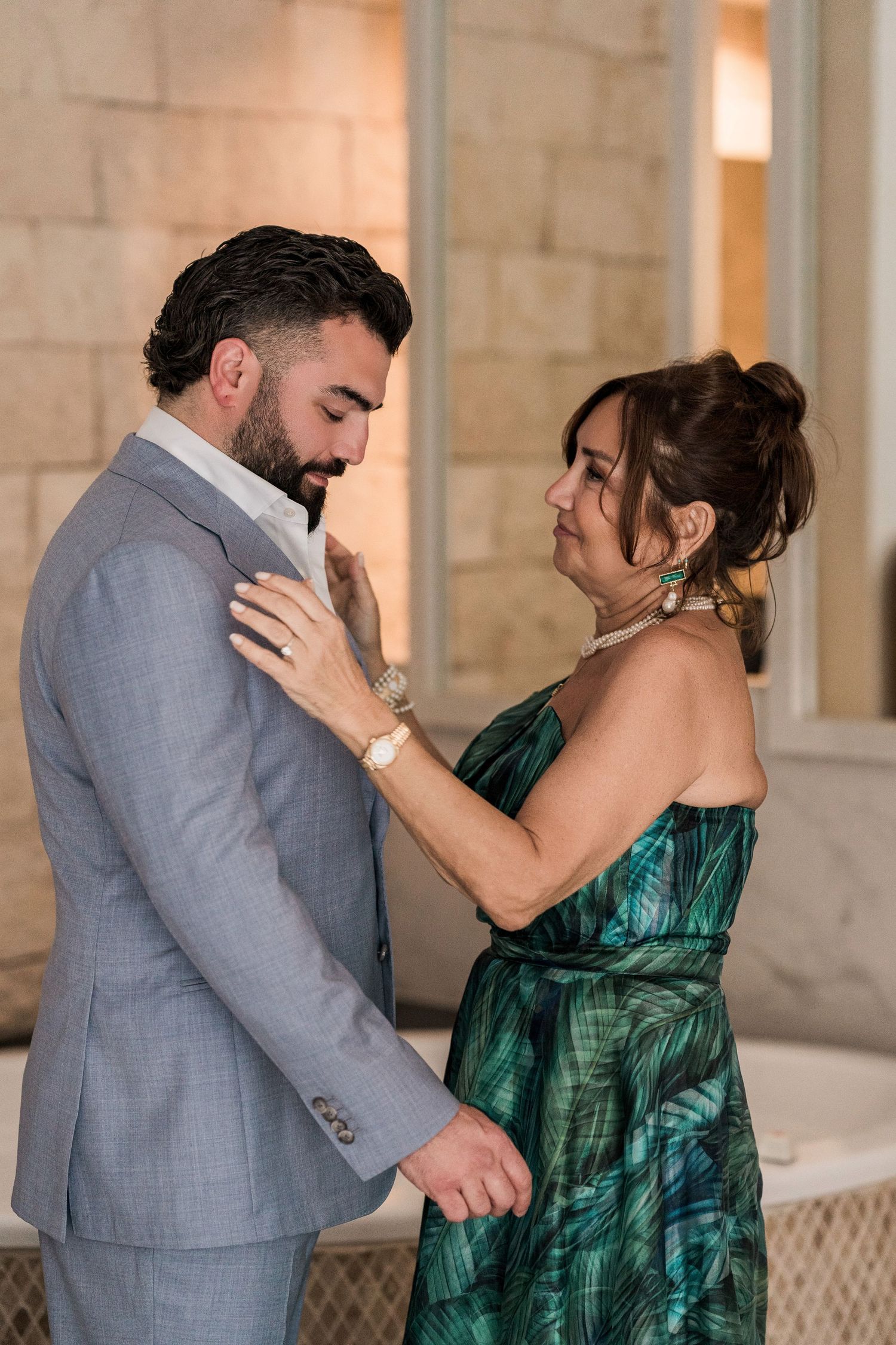 A man in a gray suit and woman in a green dress share a tender moment, with her hands resting on his chest.
