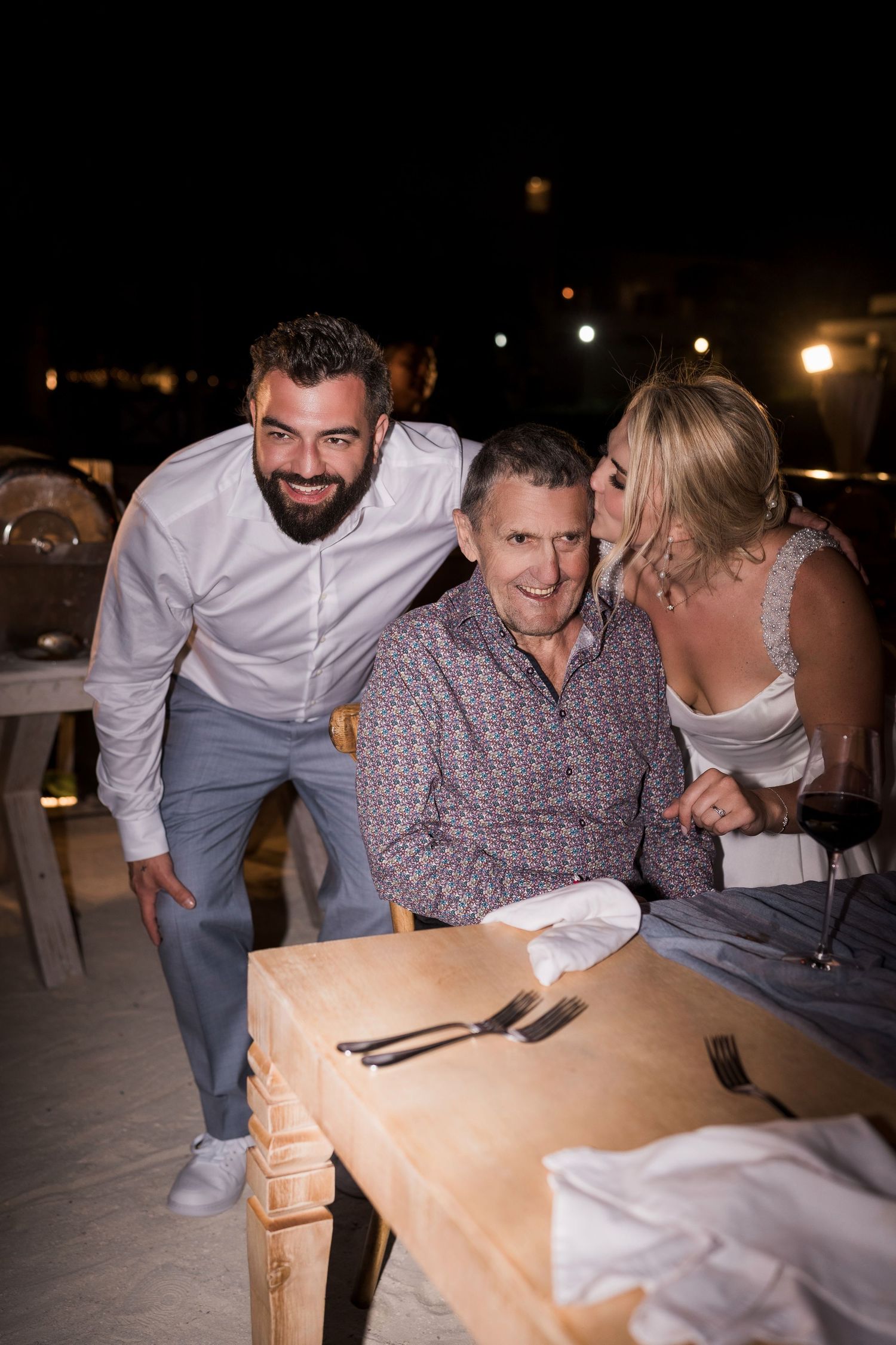 Three people posing at an outdoor dinner table at night, woman kissing older man on cheek while bearded man smiles.