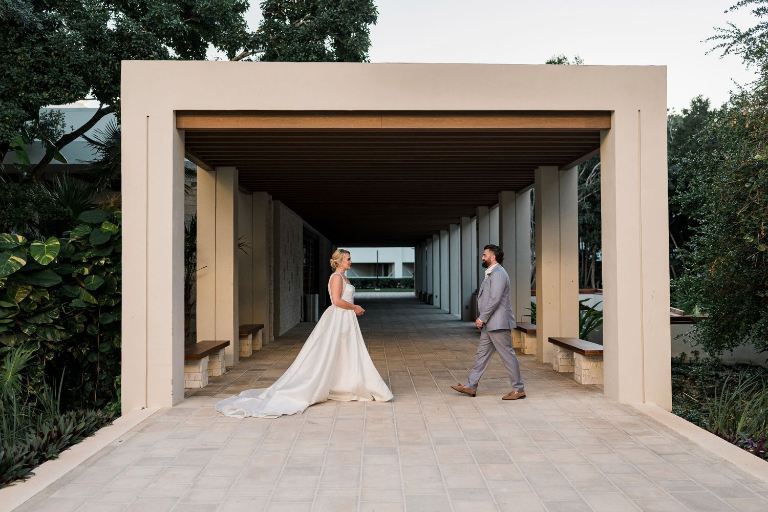 Bride and groom walk together under a modern pergola at an elegant outdoor wedding venue surrounded by lush greenery.