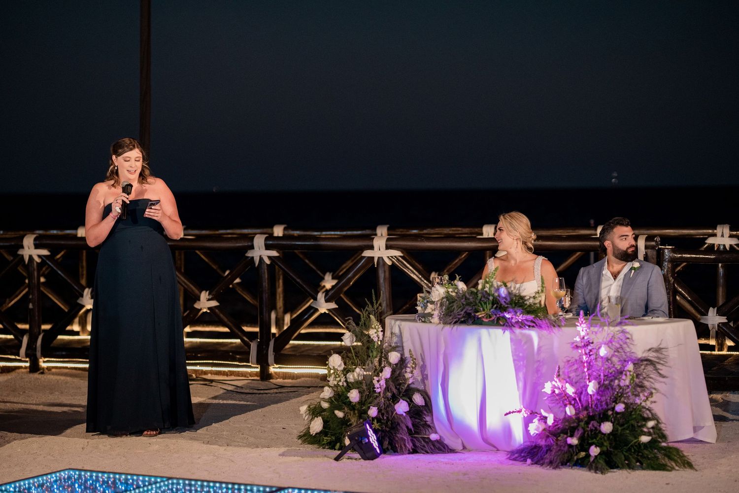 Maid of honor giving a heartfelt speech at an outdoor wedding reception while bride and groom sit at decorated sweetheart table.