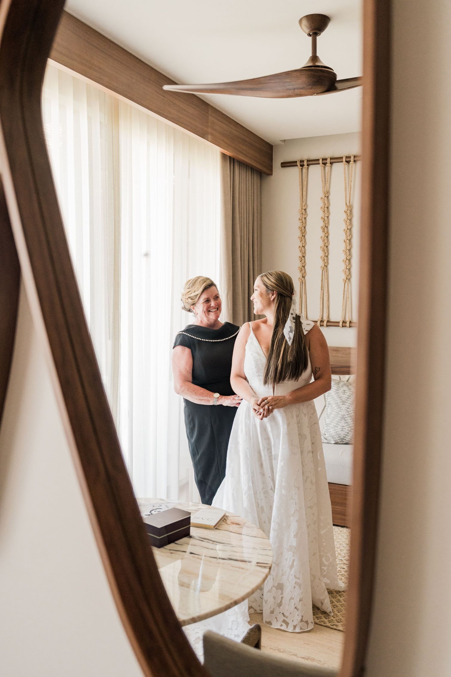 Bride in white lace gown stands with woman in black dress in elegant hotel room, reflected in ornate wooden mirror.
