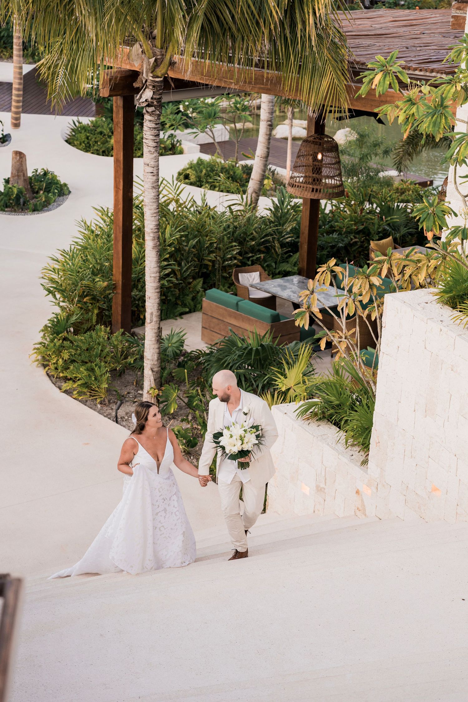 Bride and groom walk hand-in-hand through a lush tropical resort garden, bride in white lace gown holding bouquet.