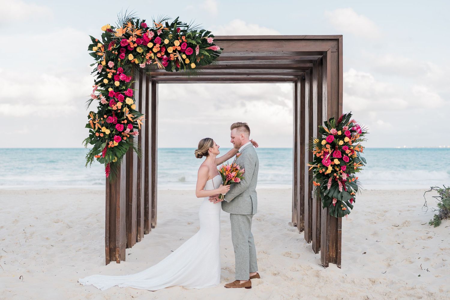 Couple exchanging vows under a floral wooden arch on a tropical beach wedding ceremony with ocean backdrop.
