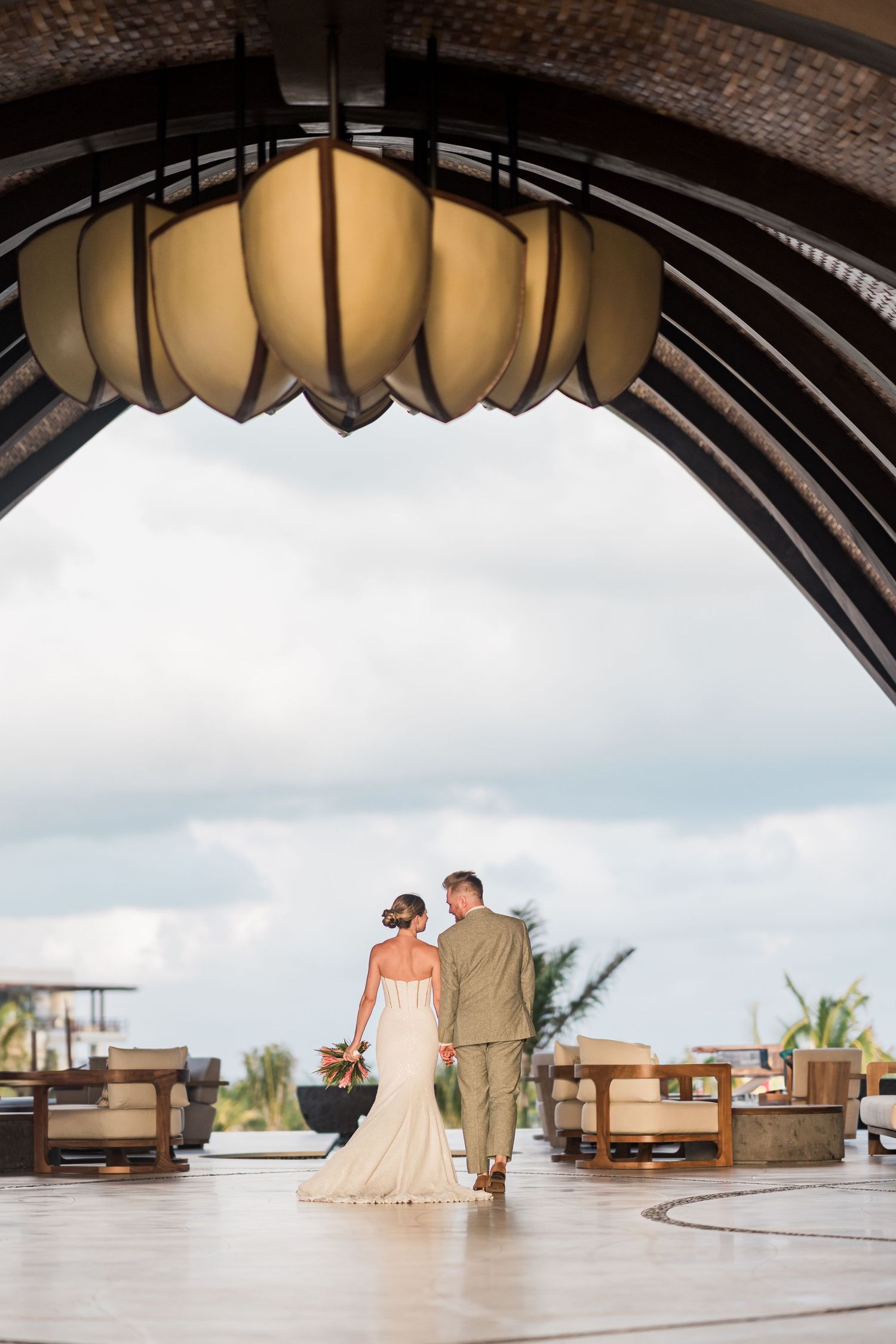 Bride and groom walk hand-in-hand under elegant arch with lotus chandelier at tropical resort wedding venue.