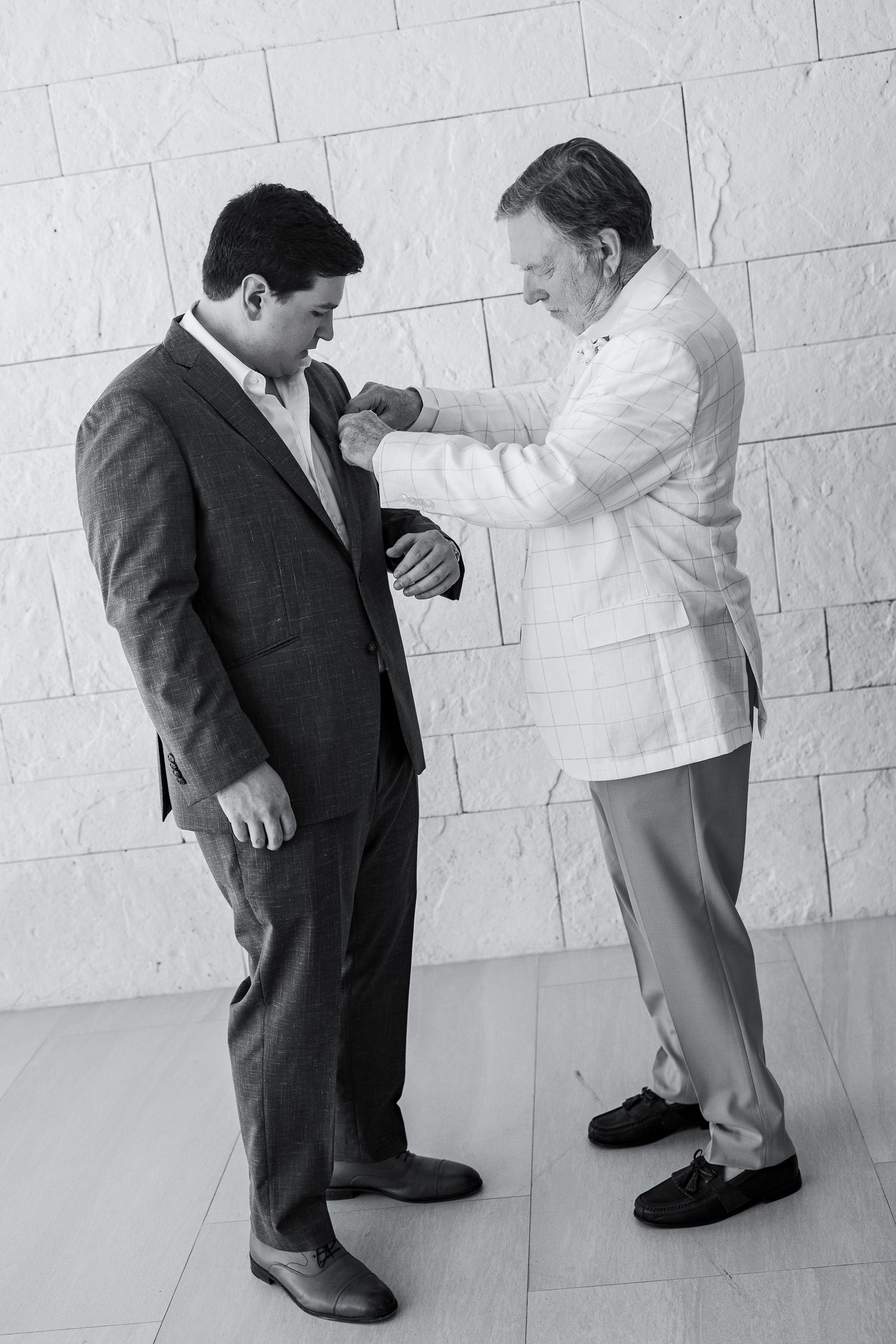 Black and white photo of two men in suits, one adjusting the other's jacket lapel near a stone wall.