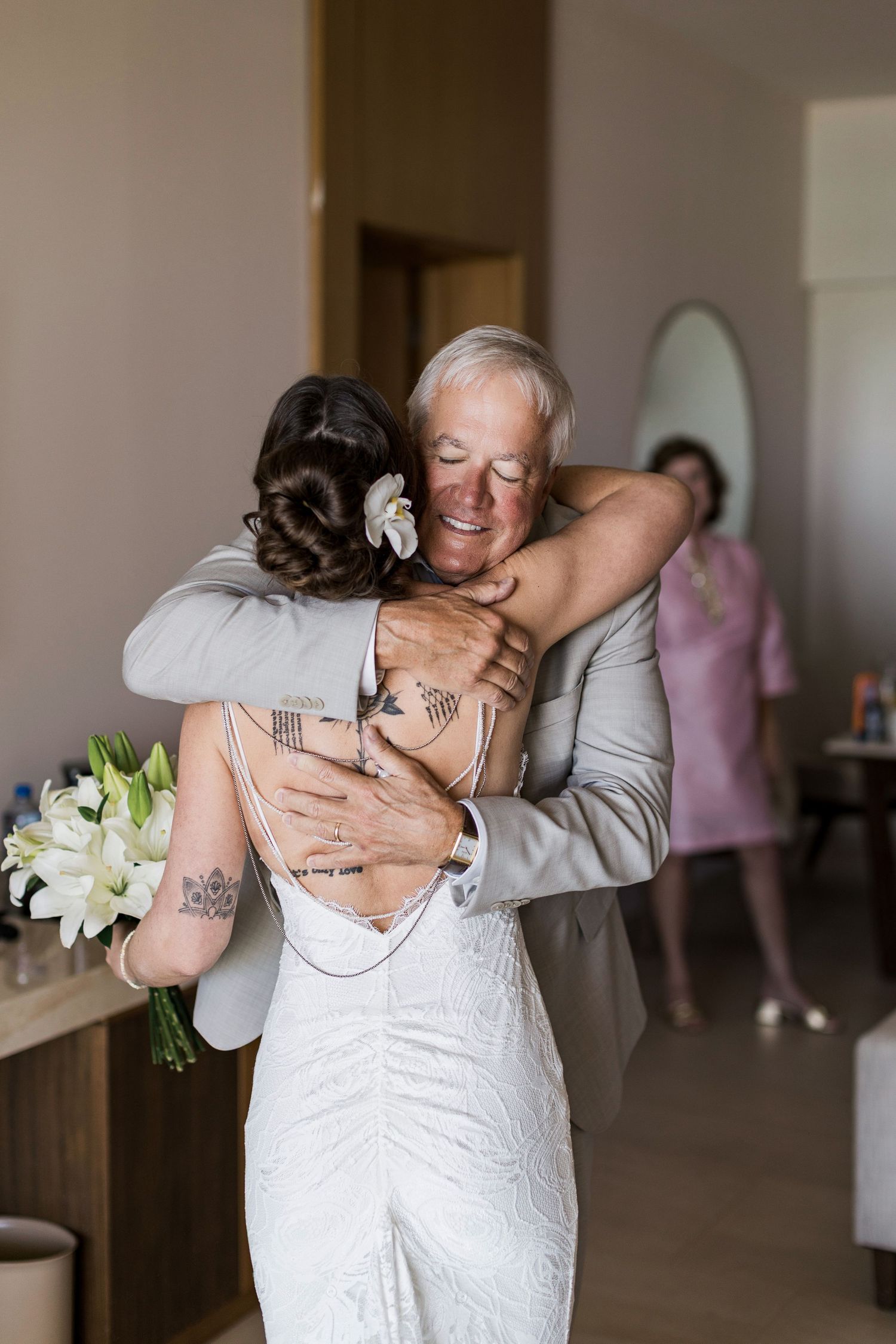 Emotional father-daughter hug at wedding, bride in white gown holding bouquet, groom smiling warmly indoors.