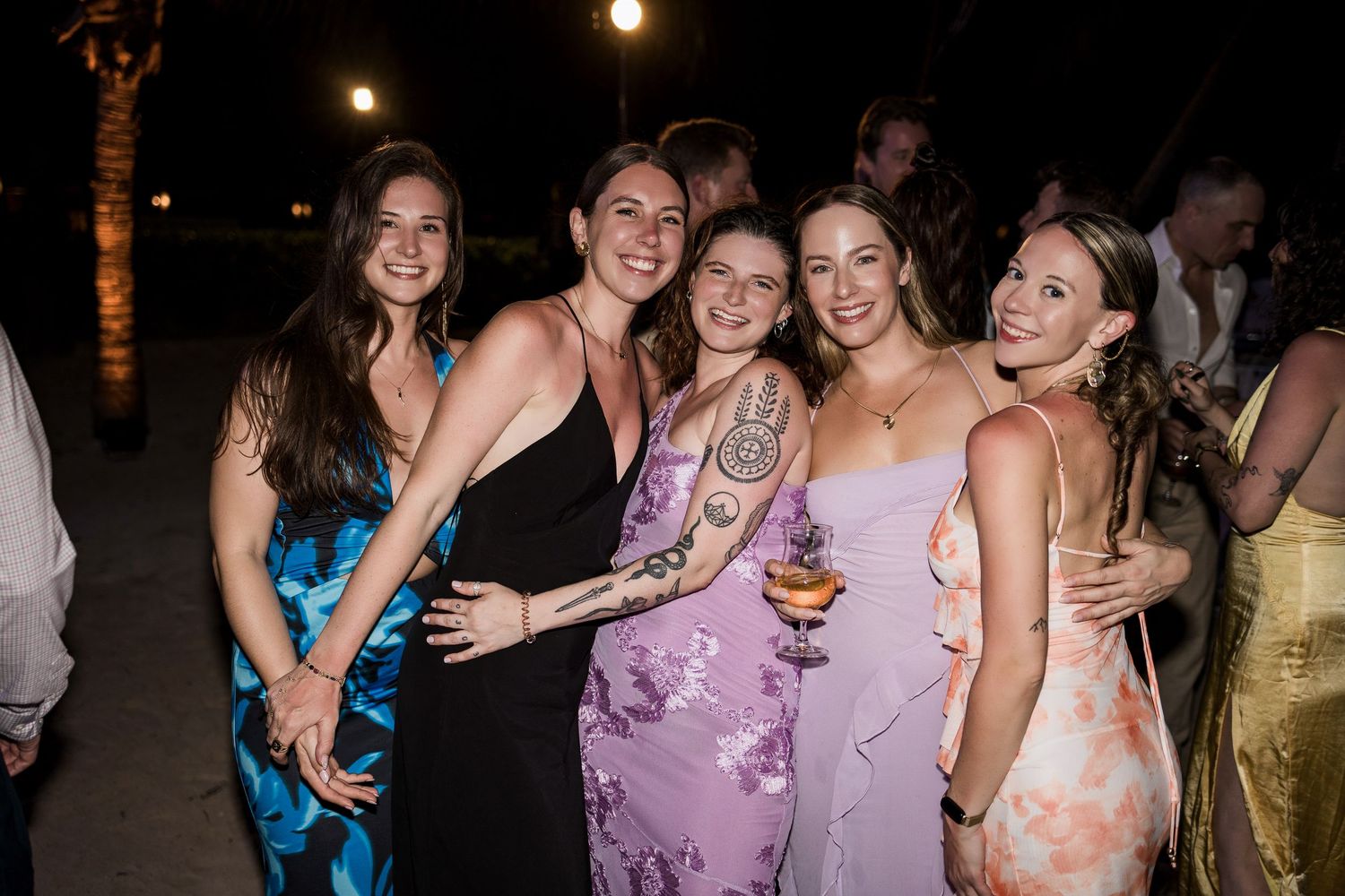 Five women in elegant dresses smile together at an outdoor evening event or wedding reception.