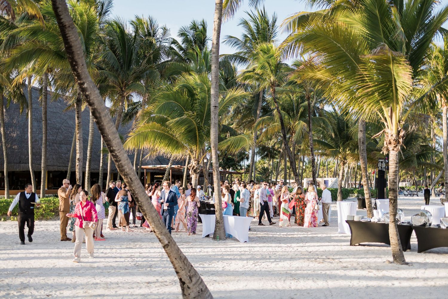 Guests mingle at an elegant outdoor beach wedding reception surrounded by tall palm trees on white sand.