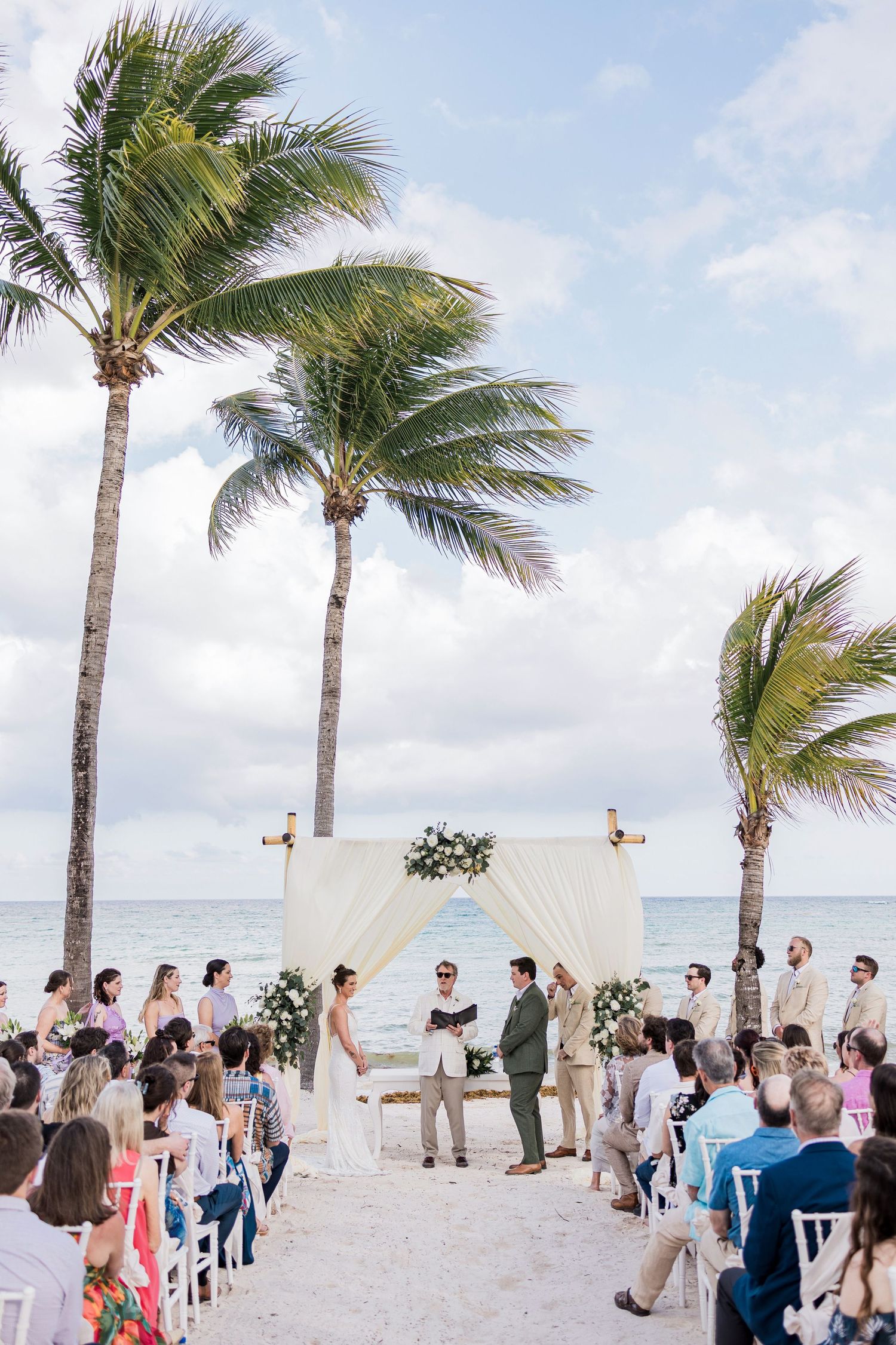 Romantic beach wedding ceremony with palm trees, white floral arch, and ocean backdrop on a sunny day.