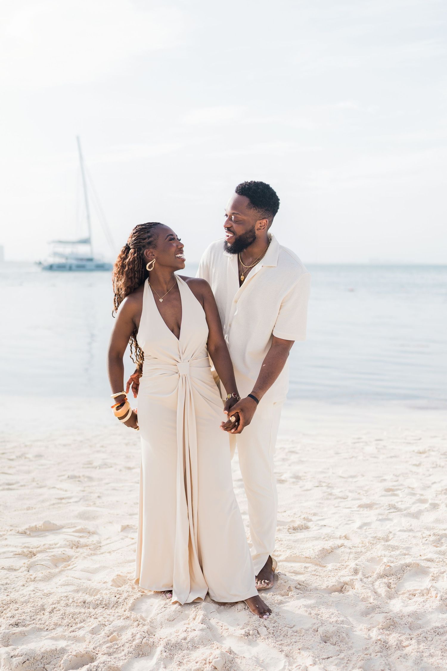 A happy Black couple in matching cream outfits hold hands on a white sand beach with a sailboat in the background.