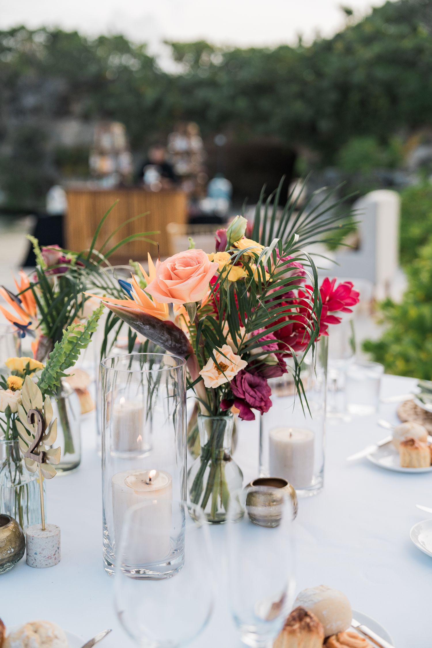 Hotel Xcaret México Tropical wedding table centerpiece with colorful flowers, palm leaves, and white candles on a white tablecloth.