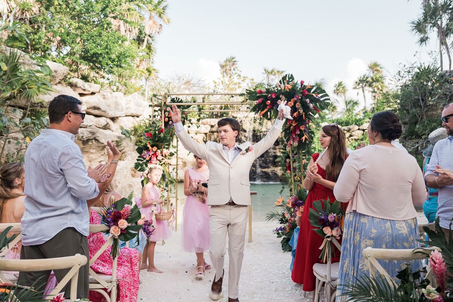 Groom celebrates with raised arms walking down the aisle at a tropical outdoor wedding ceremony with floral decor.