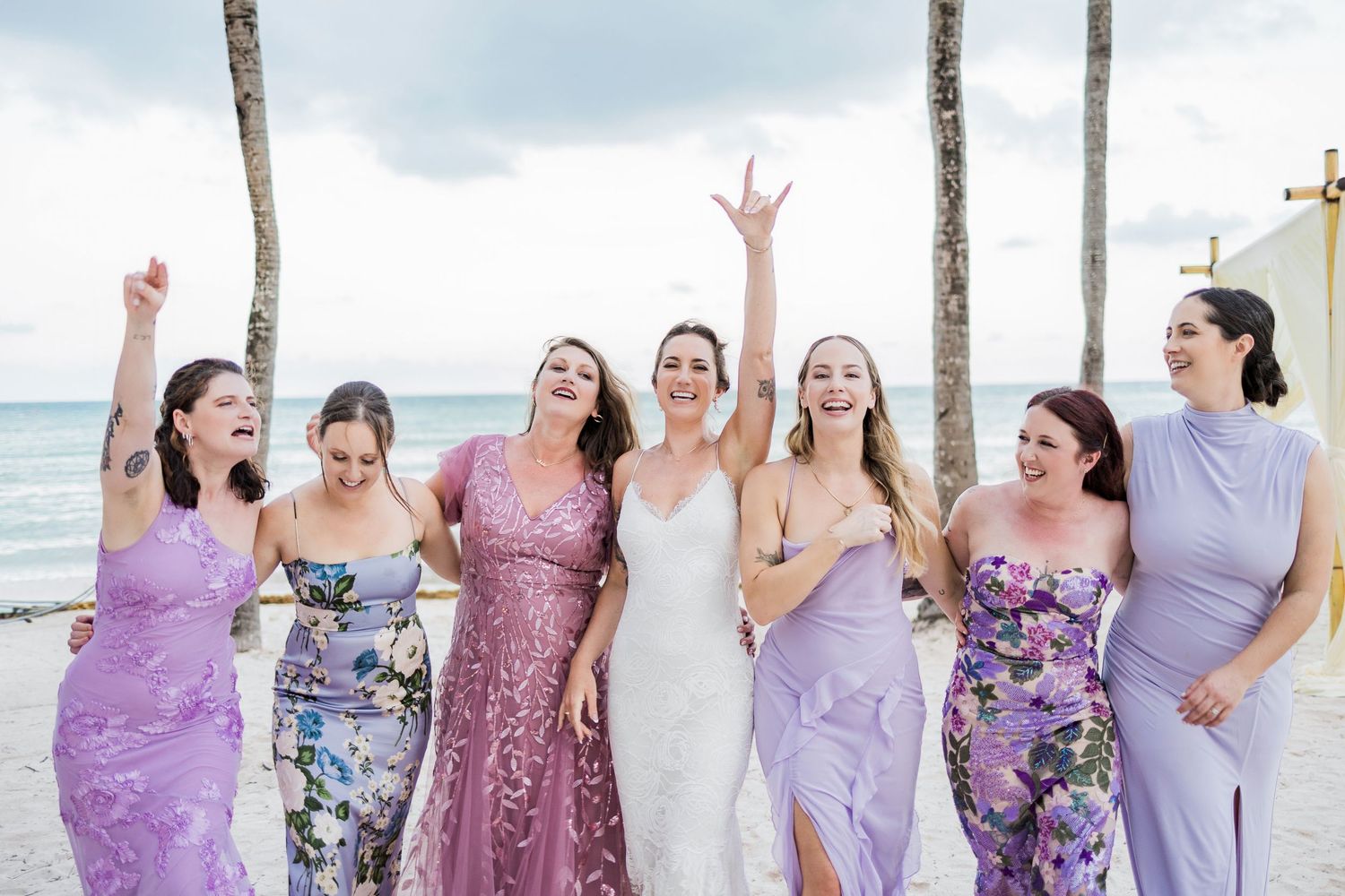 Bride in white gown celebrates with six bridesmaids in purple dresses on a beach, all laughing and raising arms.
